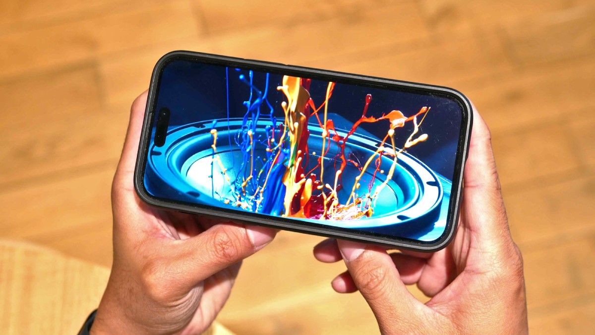 A person holds a smartphone displaying a vivid splash of colored paint above a speaker, set against a wooden background.
