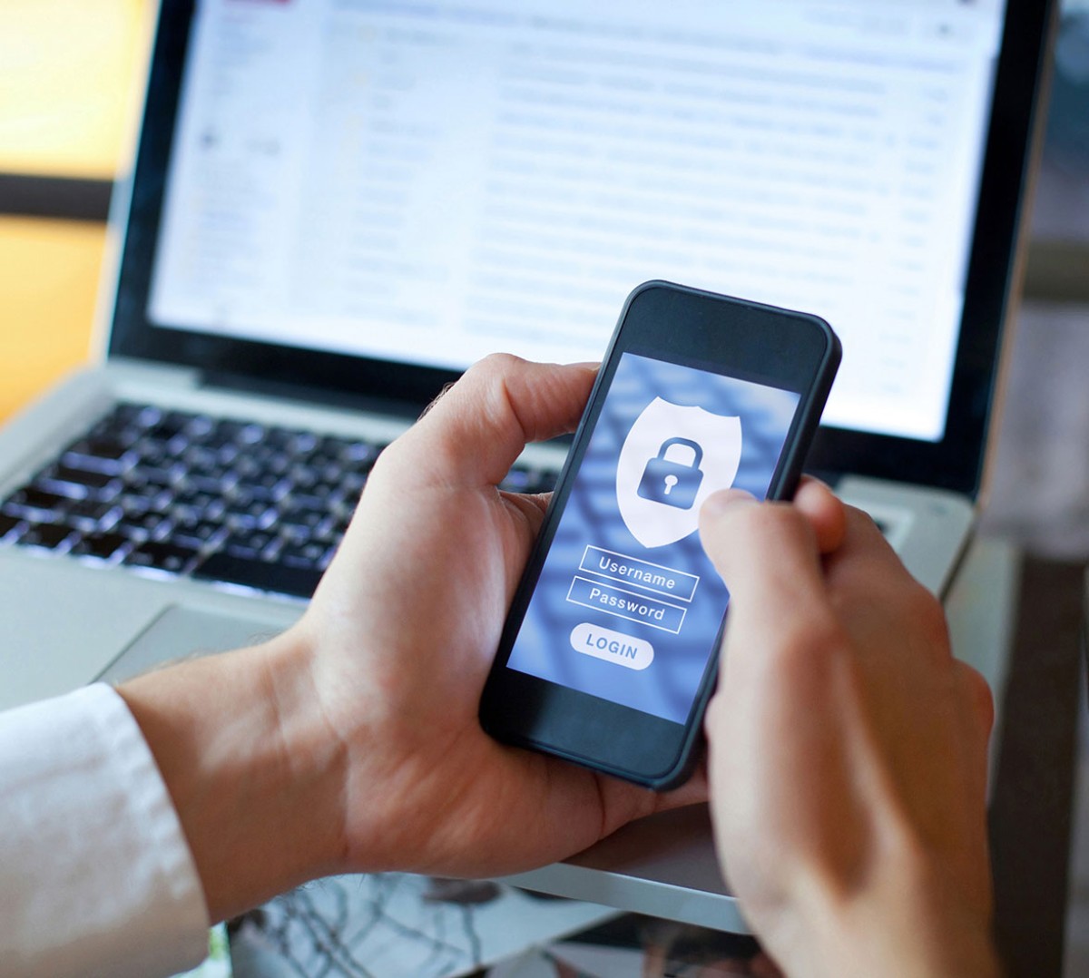 A person holding a smartphone displaying a login screen with a padlock, while a laptop is open in the background.