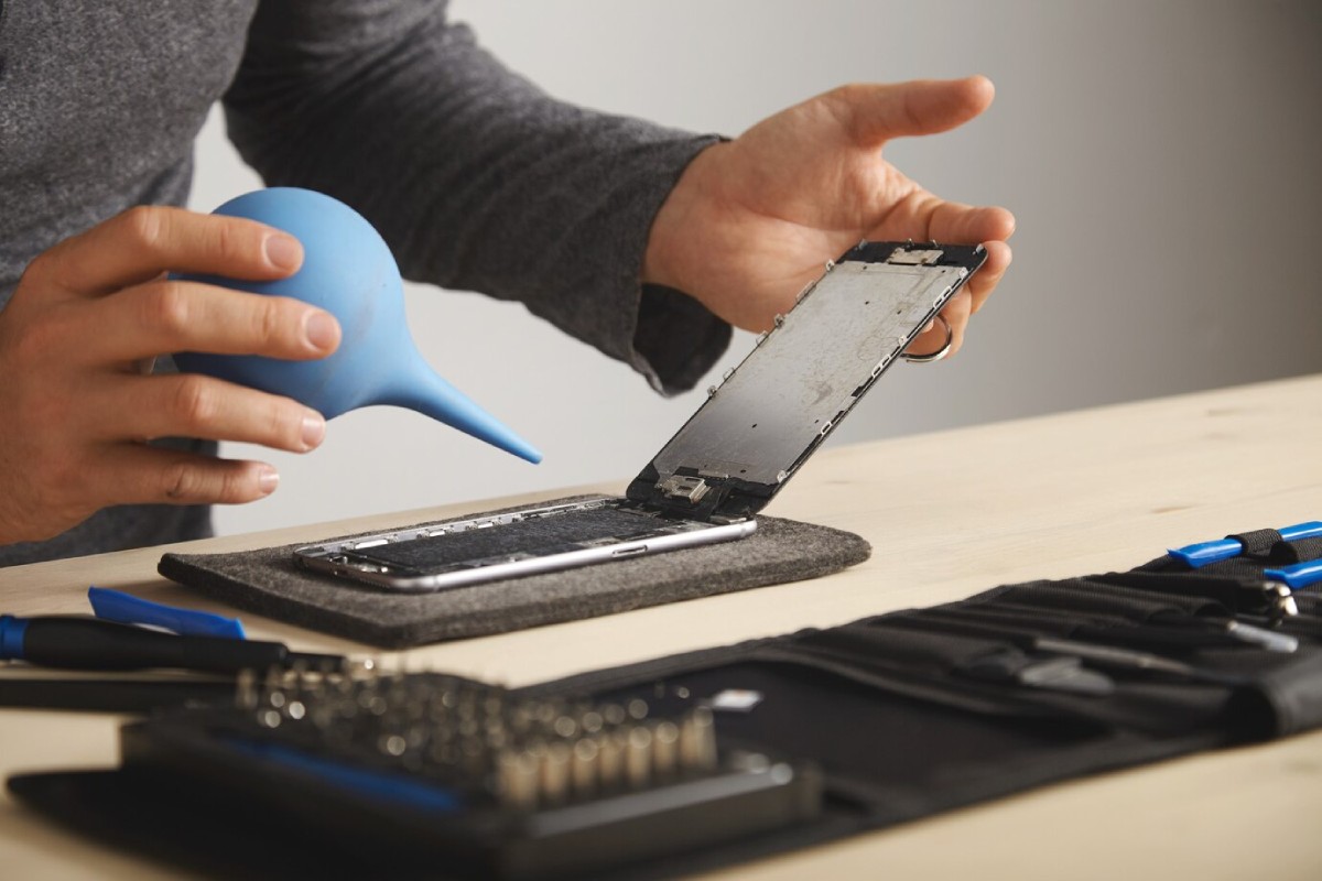 A person disassembles a smartphone, using a blue air blower, with tools neatly organized on a black tool roll.