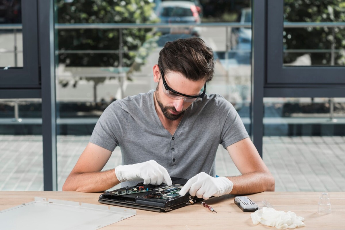 A person in a gray shirt repairs a device, wearing white gloves, with tools and a clean workspace in bright, modern surroundings.