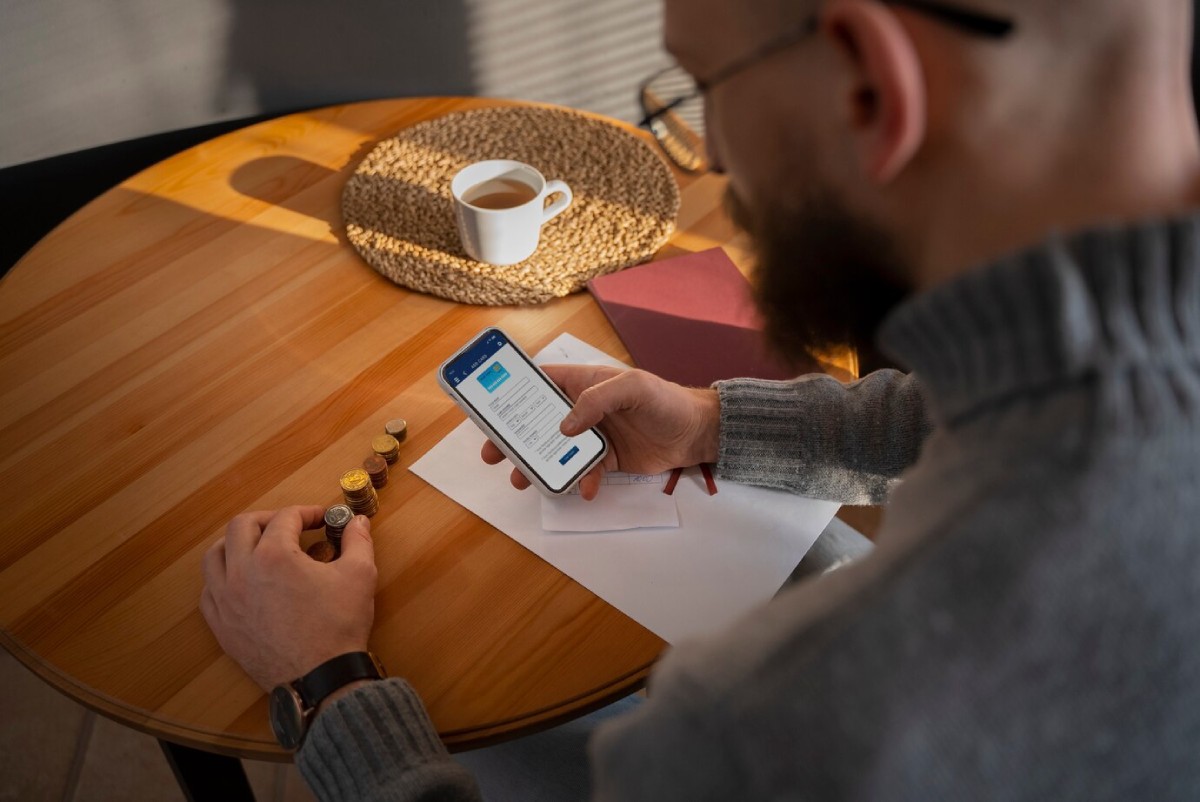 A person sits at a wooden table, checking a mobile phone with notes, coins, and a cup of coffee nearby.