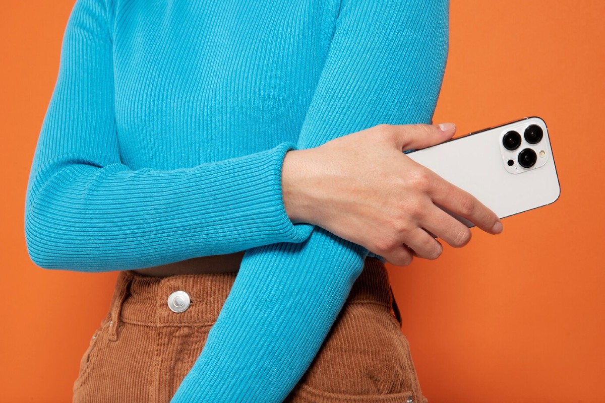 A person in a blue ribbed sweater holds a white smartphone against an orange background, adding a pop of color to the scene.