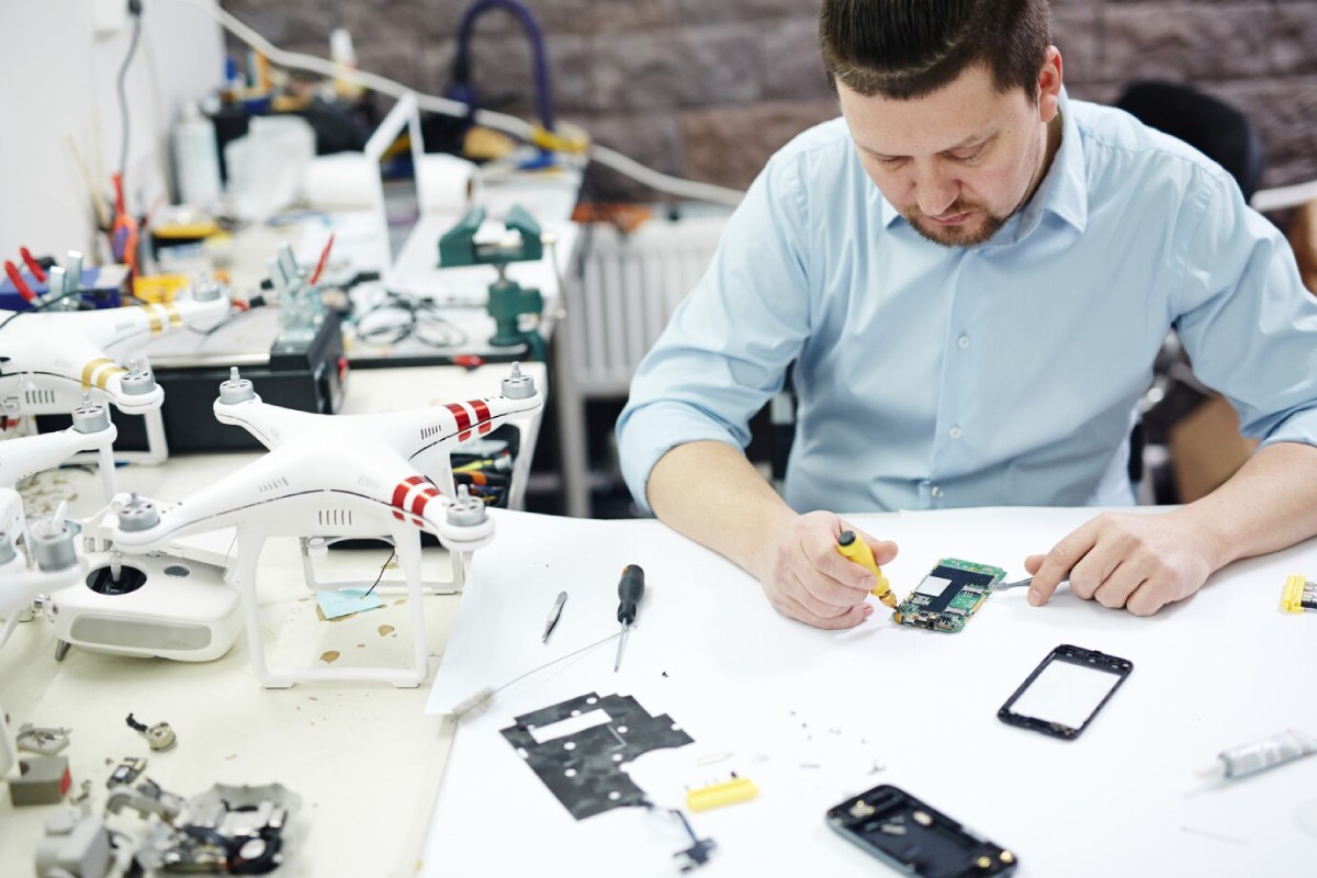 A technician repairs a smartphone on a cluttered workbench surrounded by drones and tools, focused on a circuit board.