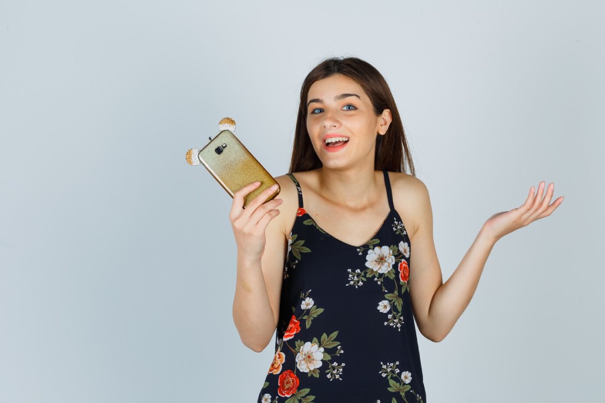 A woman in a floral dress holds a phone with a bear-themed case, gesturing with her free hand against a plain background.