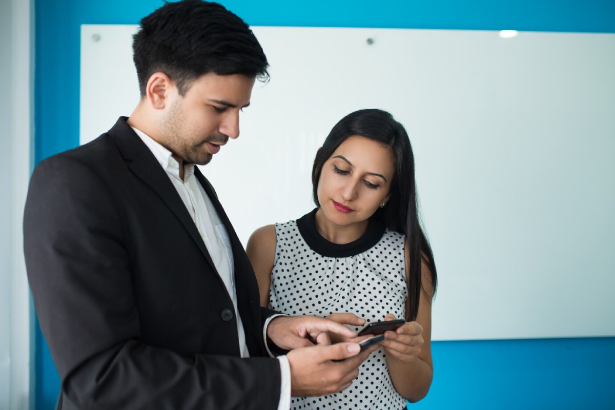 Two professionals engage in a discussion while looking at a smartphone in a modern office setting with a blue wall.