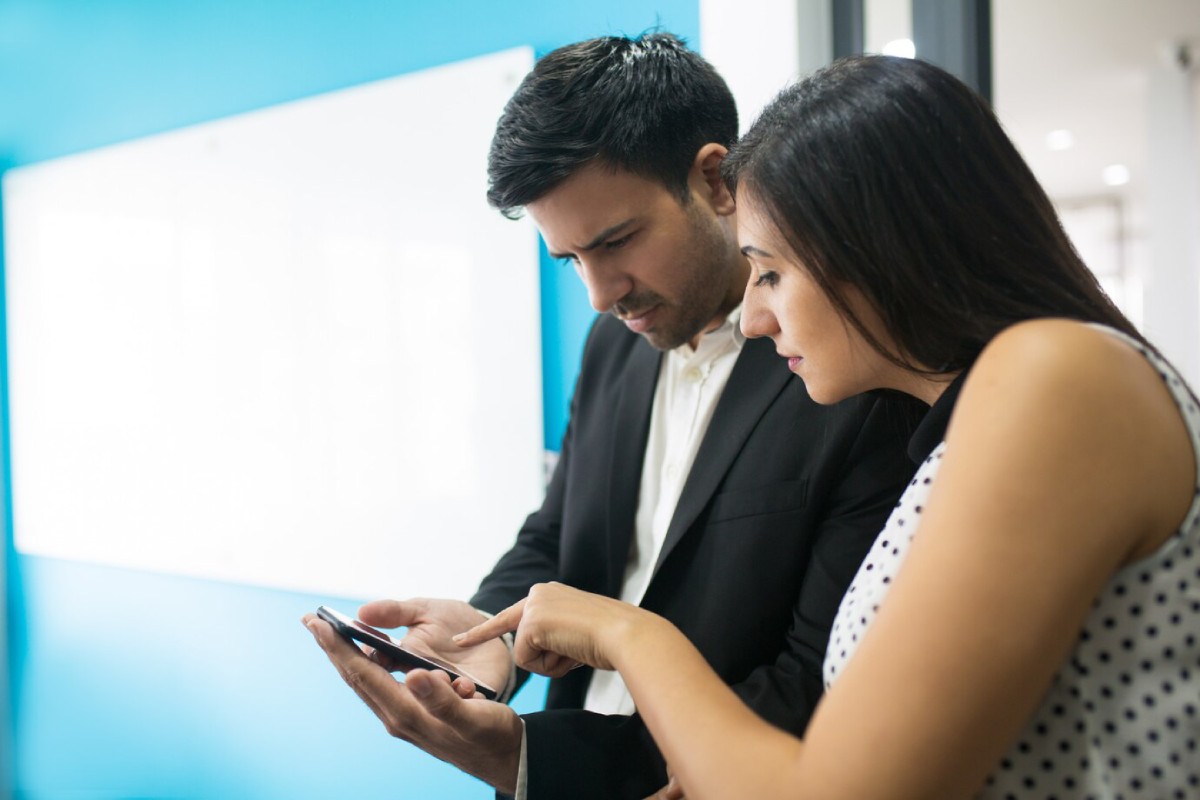 Two individuals closely examine a smartphone, engaged in conversation, in a modern office setting with a blue wall.