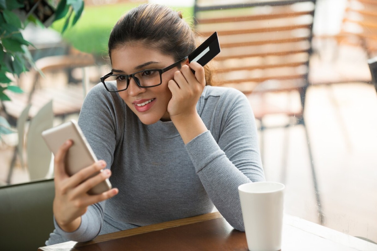 A person sitting at a table in a café, holding a smartphone in one hand and a credit card in the other, looking pensive.