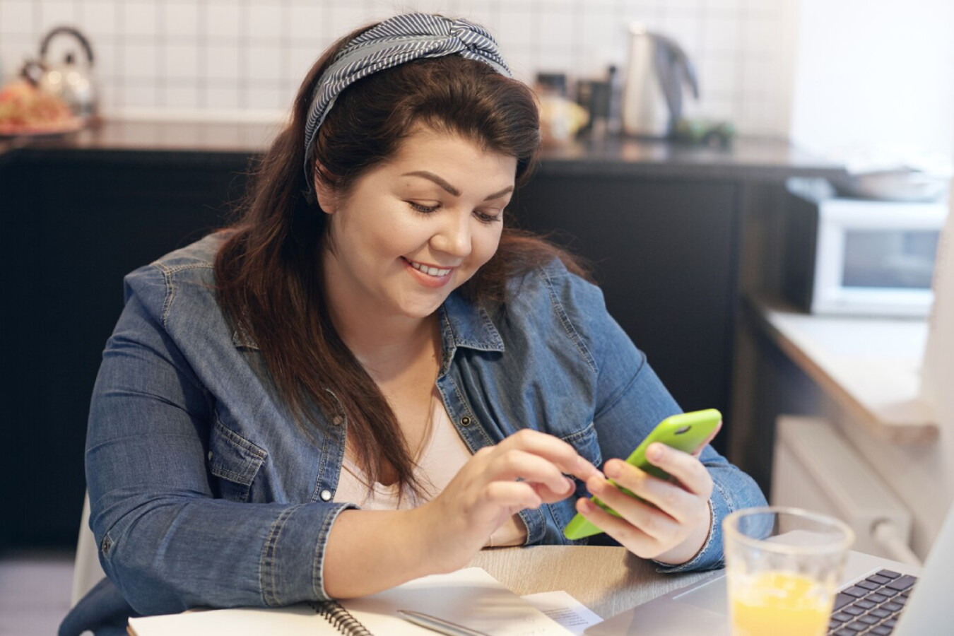 A woman in a denim shirt and headband sits at a table, using a green smartphone, with a glass of juice and a notepad nearby.