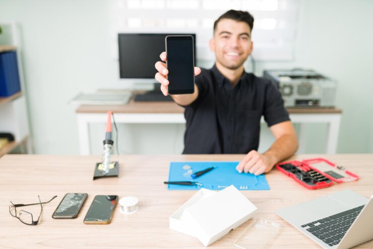A technician presents a smartphone while seated at a desk with tools, glasses, and a laptop in a workspace setting.