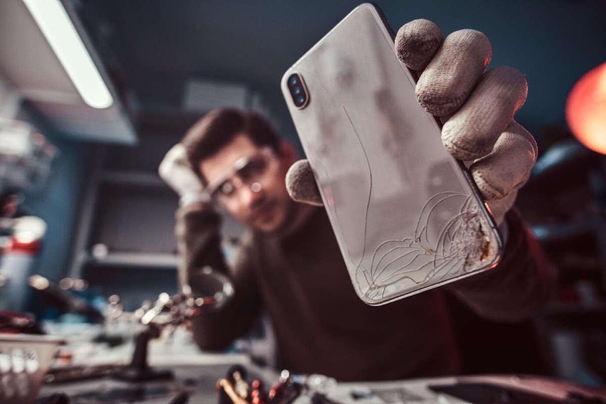 A person in gloves holds a damaged smartphone with visible cracks, surrounded by tools in a dimly lit workspace.