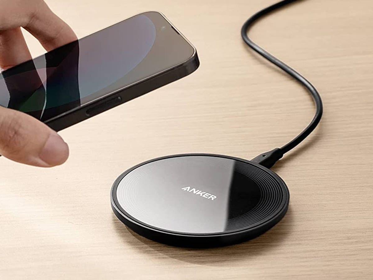 A person holds a smartphone above a sleek black Anker wireless charger on a wooden surface, ready to charge it.