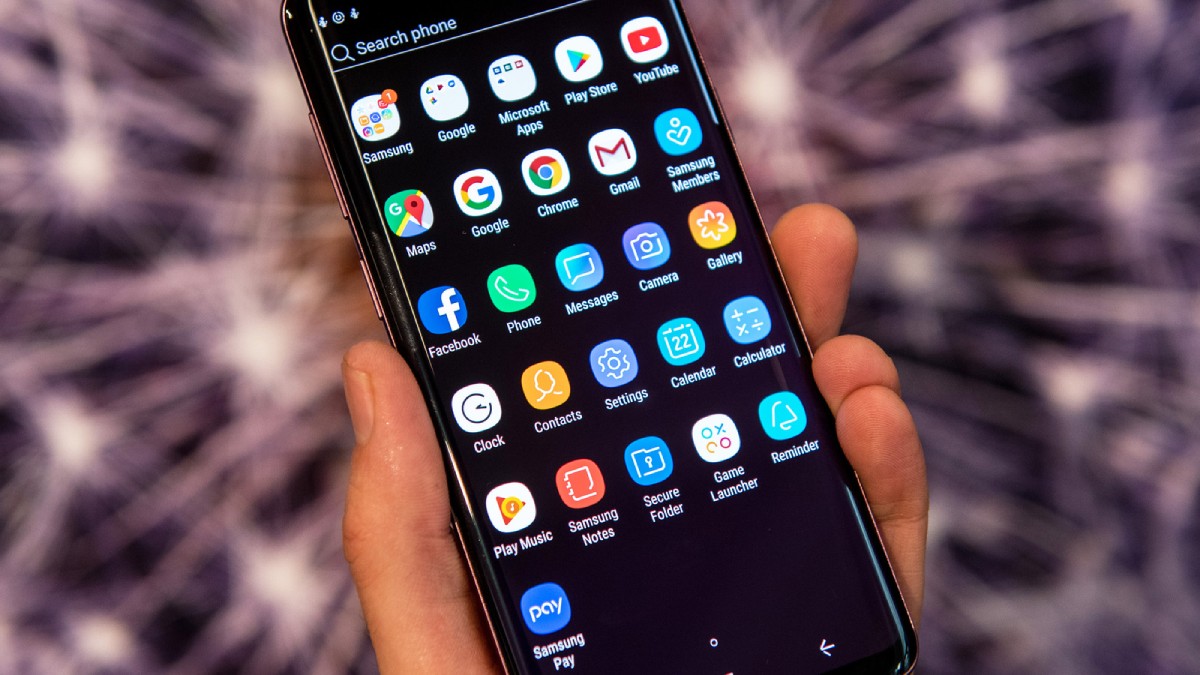 A person's hand holds a smartphone displaying a grid of app icons, including social media and productivity apps, on a dark background.
