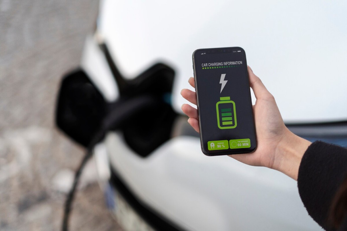 A hand holds a smartphone displaying car charging status at 90%, with a charging icon, next to an electric vehicle charging port.