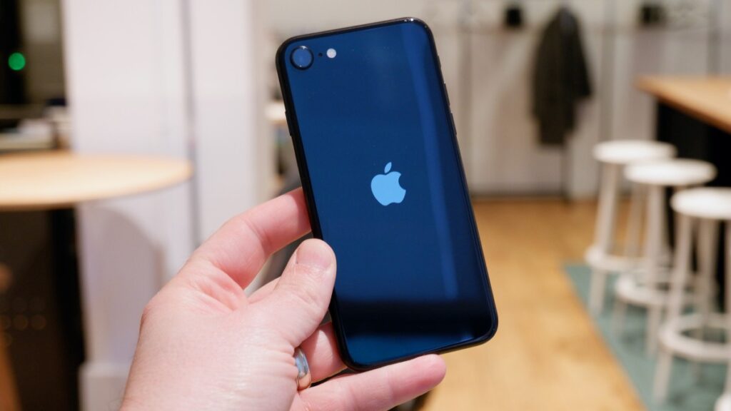 A person holds an iPhone SE with a glossy blue back displaying the Apple logo, set against a blurred interior background.