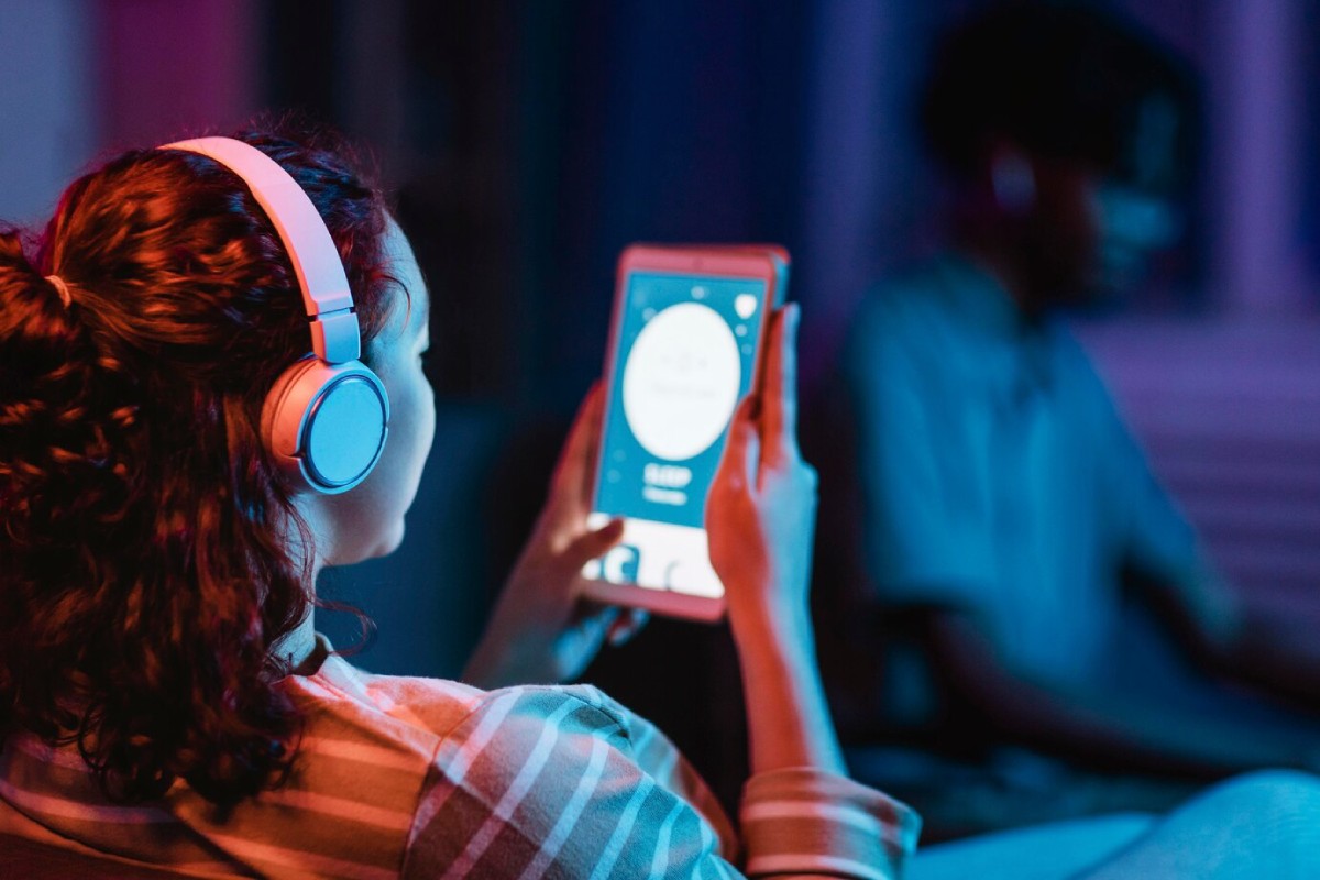 A woman with curly hair wearing pink headphones holds a tablet, displaying a music app, amidst a softly lit, colorful background.