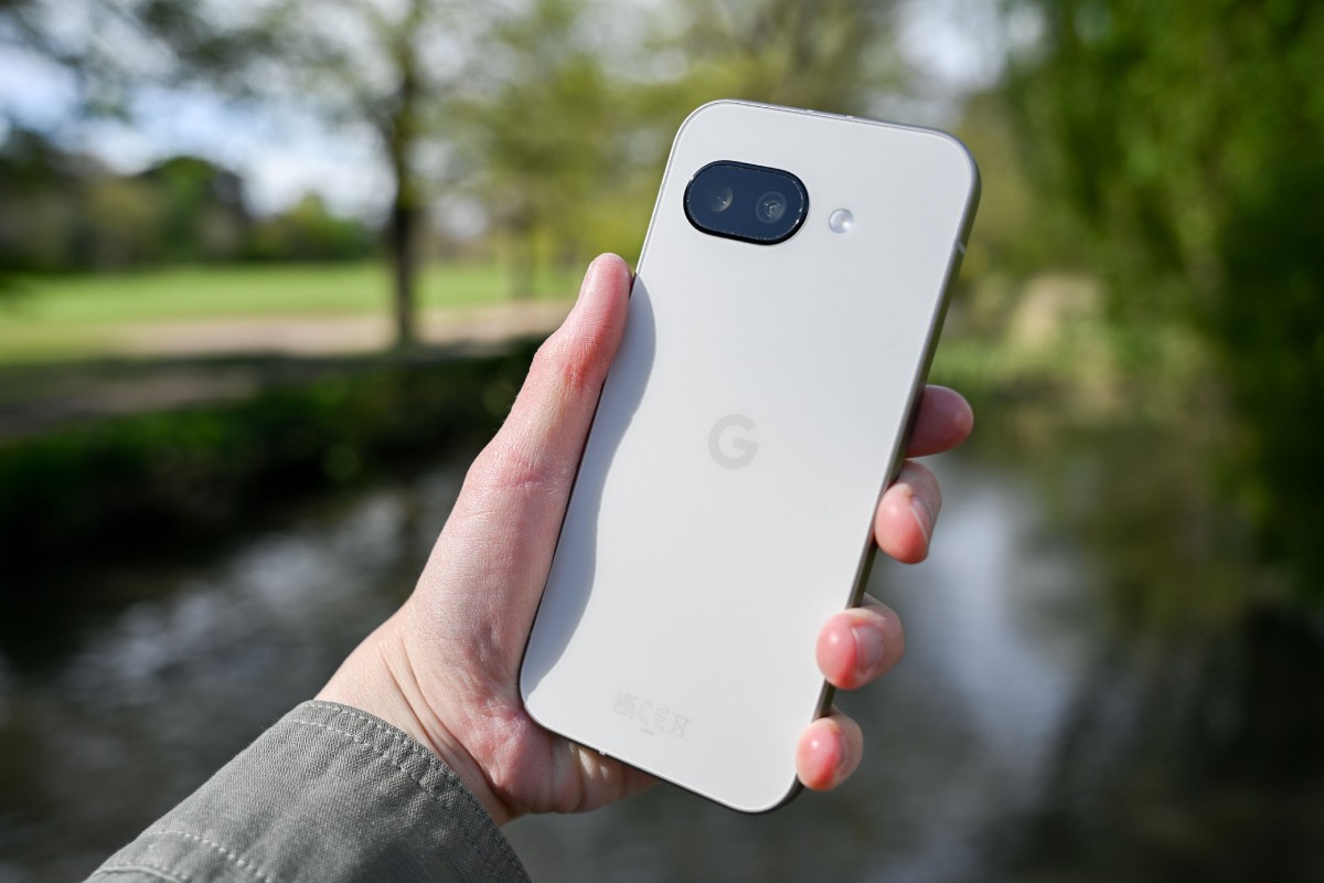 A person holds a white Google smartphone outdoors by a stream, with blurred greenery in the background.