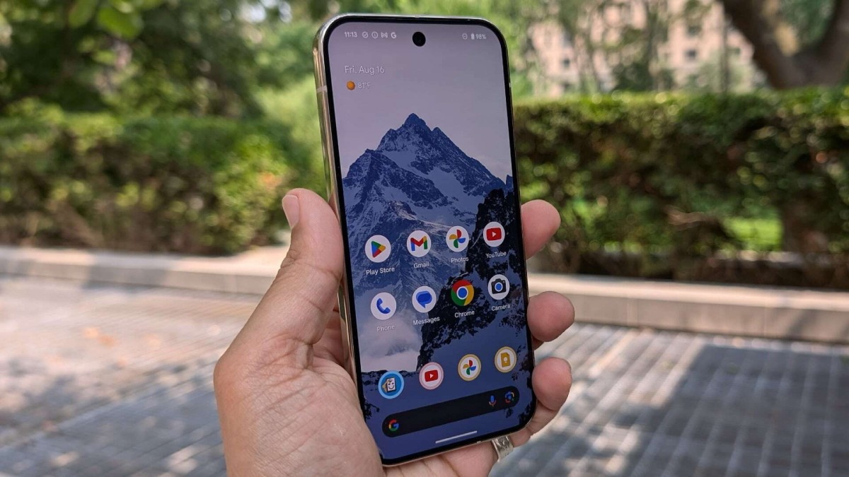 A person holding a smartphone outdoors displaying a mountain wallpaper and various app icons on the screen.