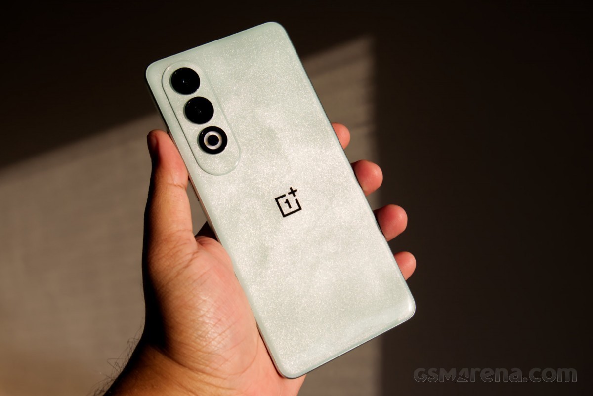 A person's hand holding a mint green OnePlus smartphone with three cameras and a logo, illuminated by soft natural light.
