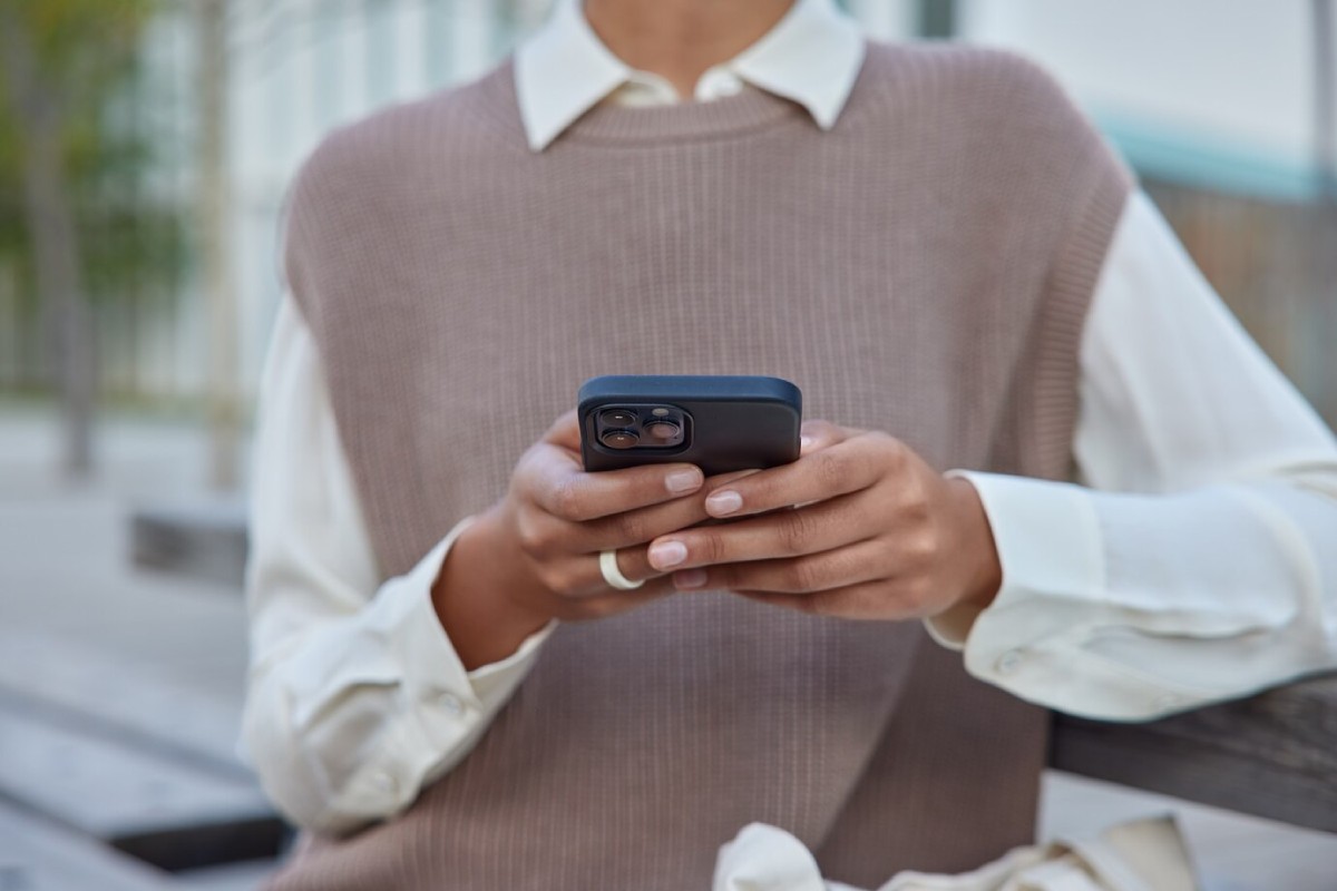 A person sits on a bench, wearing a beige sweater over a white shirt, holding a smartphone with both hands.