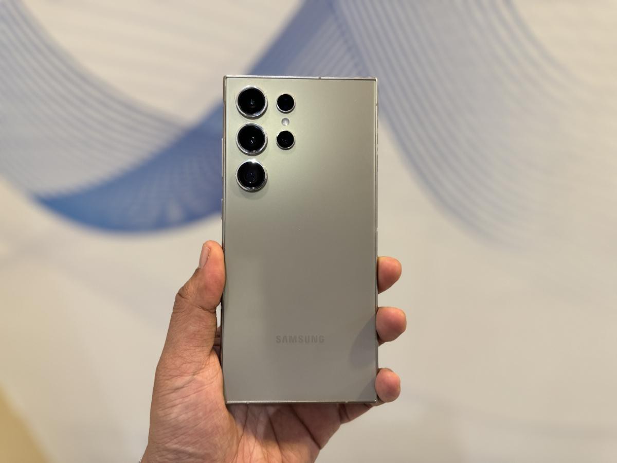 A person holds a sleek silver Samsung smartphone showing a triple-camera setup on the back against a soft, blurred background.