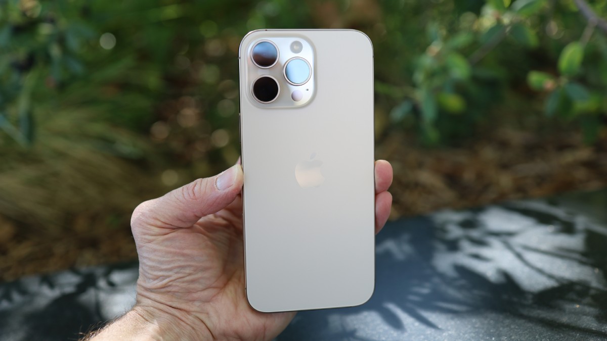 A person holding the back of a gold iPhone 13 Pro, showcasing its camera module and Apple logo against a blurred natural background.