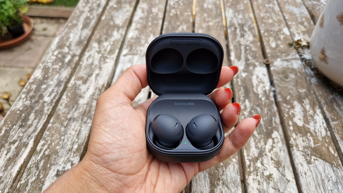 A hand holding a black wireless earbuds case, slightly open to reveal two earbuds inside, set atop a rustic wooden table.