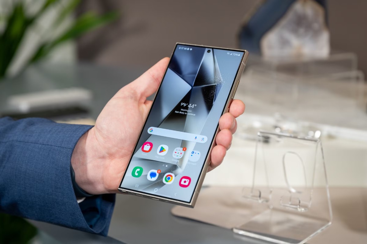 A person in a suit holds a sleek smartphone displaying app icons and the weather, set against a blurred background.