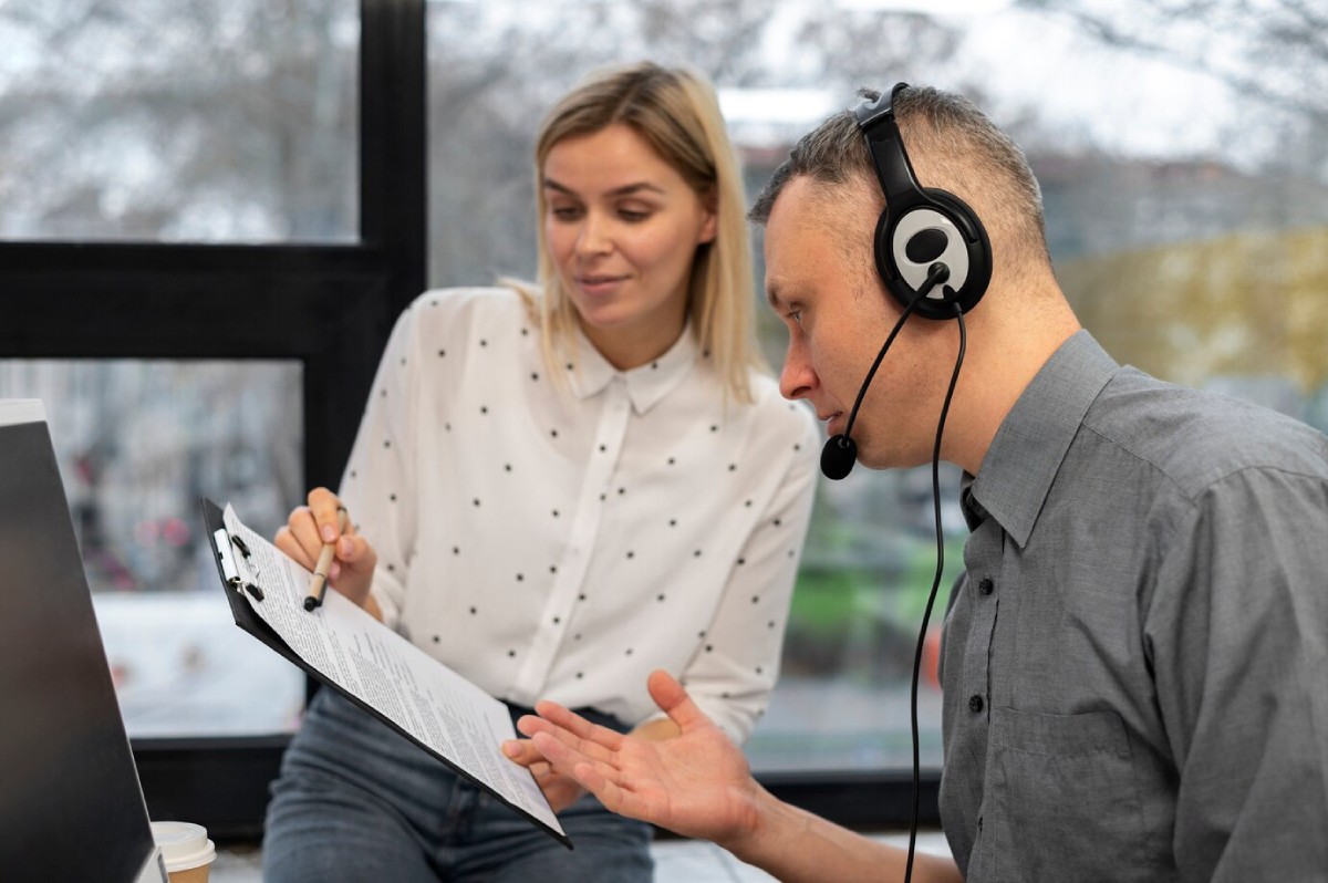 A person wearing headphones gestures while discussing a document with another individual in a bright, modern workspace.