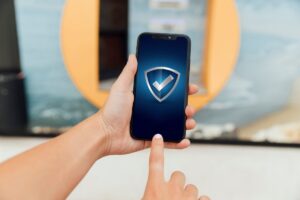 A person holds a smartphone displaying a shield icon with a checkmark, emphasizing security or verification. Background features blurred objects.