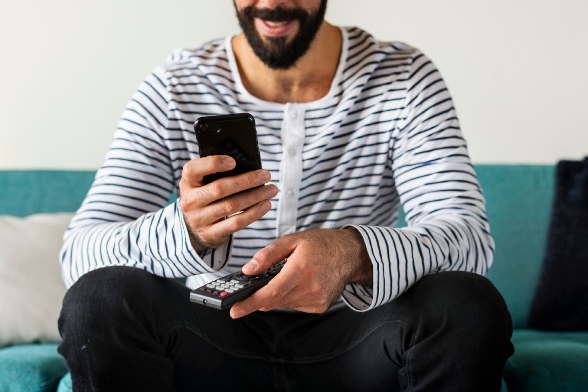 A smiling man in a striped shirt sits on a turquoise couch, holding a smartphone in one hand and a remote control in the other.