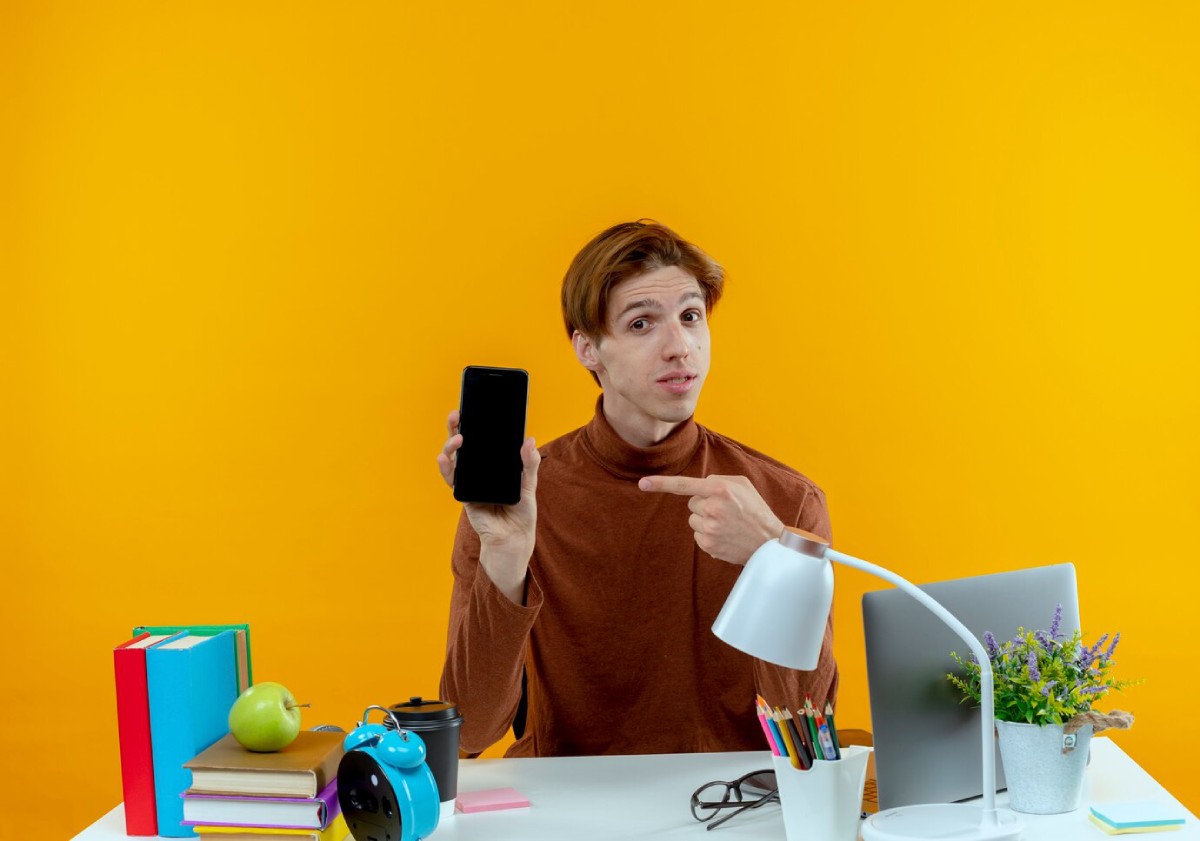 A person in a brown sweater points to a smartphone at a desk with books, a cup, a clock, and a potted plant against a yellow background.