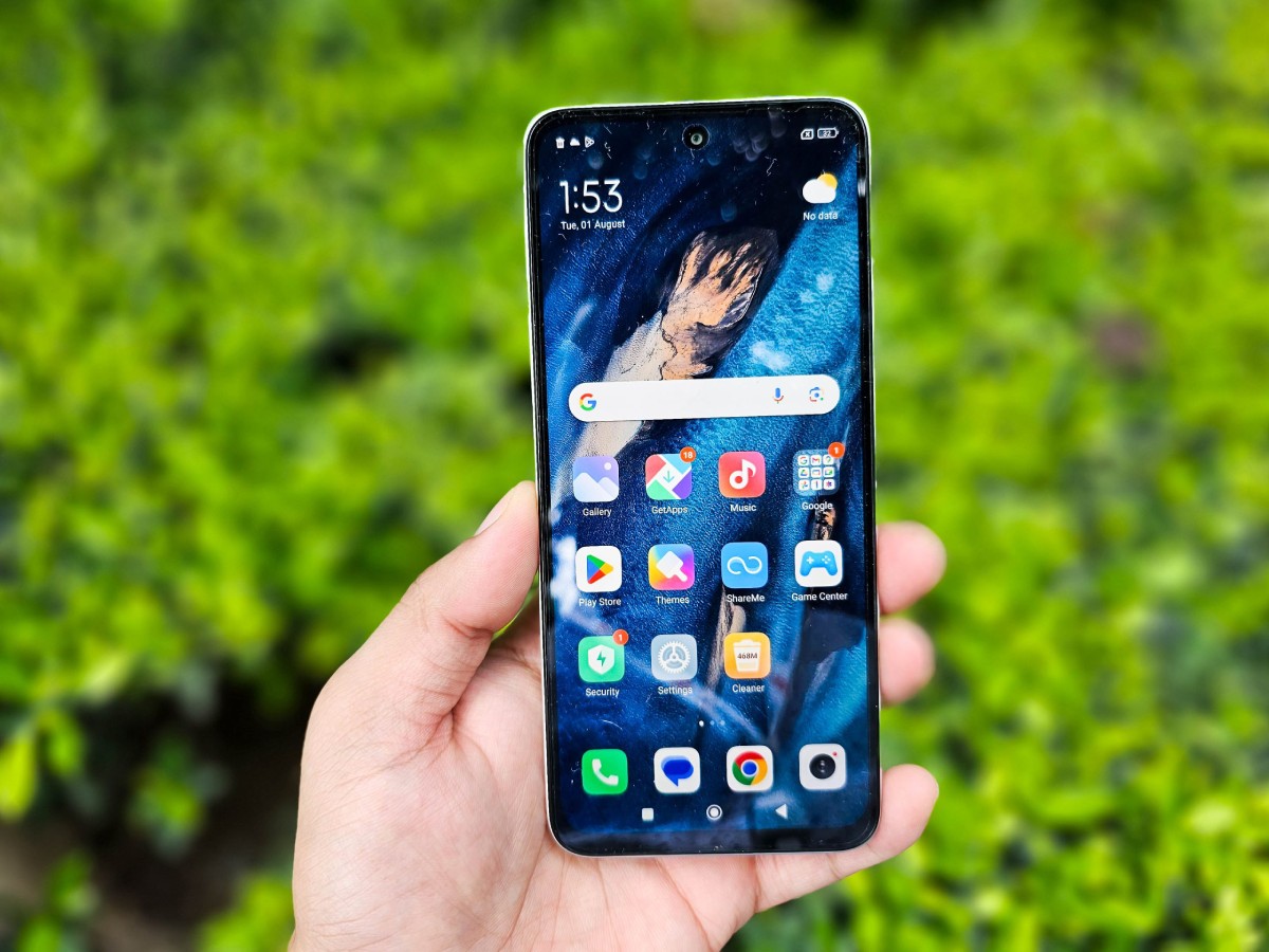A hand holds a smartphone displaying a vibrant blue wallpaper, surrounded by lush green foliage in the background. Time reads 1:53 PM.