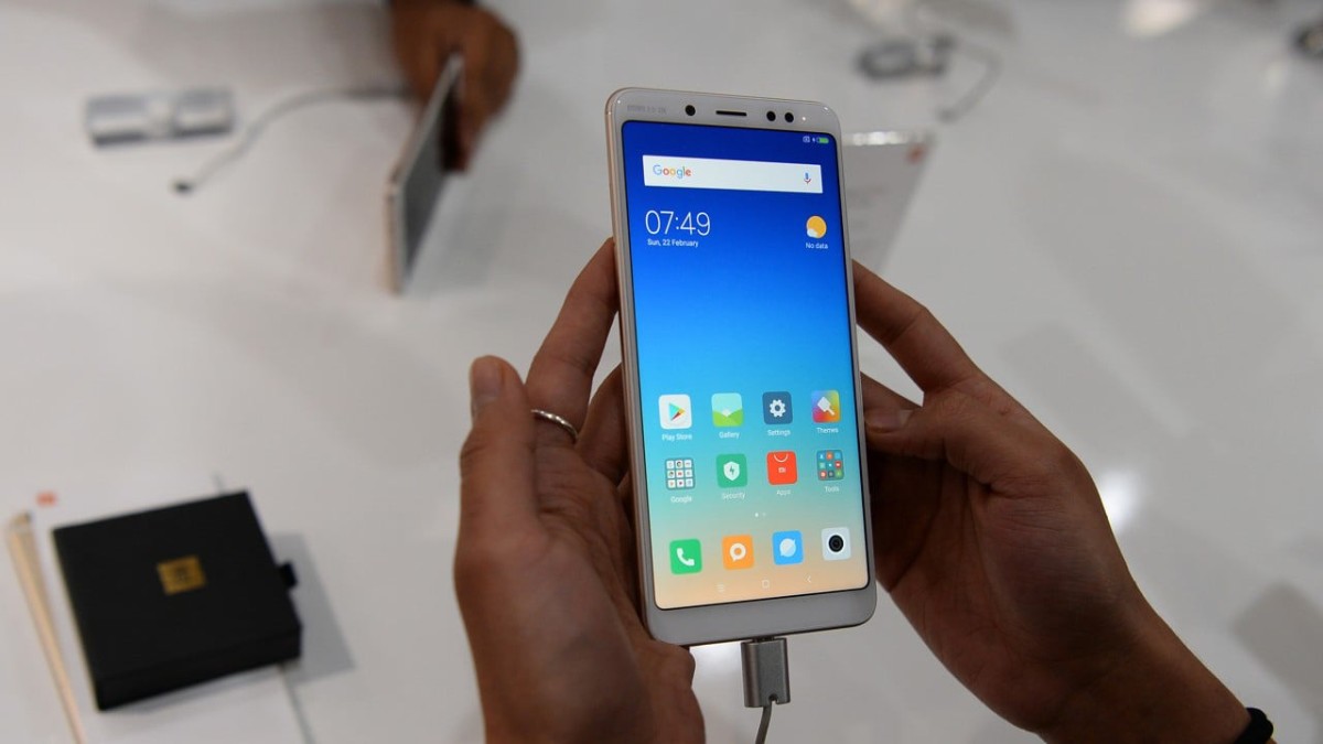A person holding a smartphone displaying the home screen with app icons, time, and date. The phone is connected to a charger.
