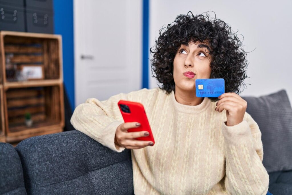 A person holding a blue credit card in one hand and a red smartphone in the other, seated on a gray couch in a cozy room.